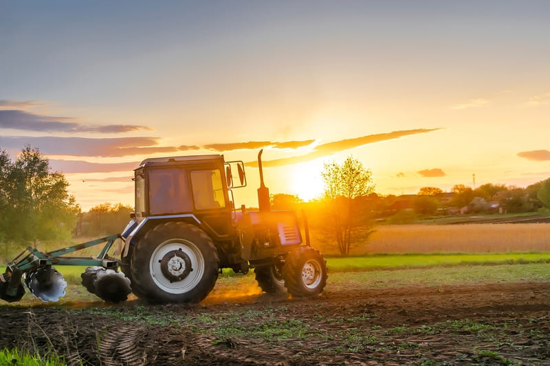 A blue farm tractor plowing a field during a vibrant sunset, casting long shadows across the landscape.