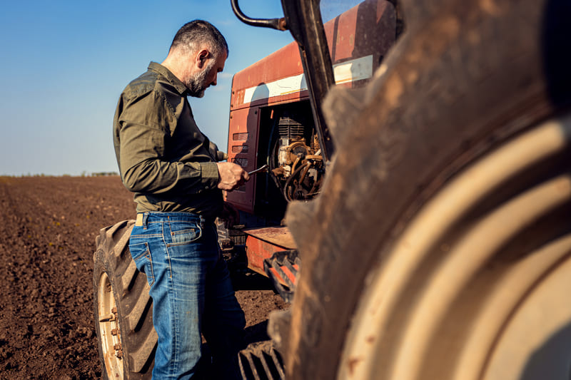 A farmer in a work hat and overalls is repairing a large tractor in an open field, using tools to adjust engine components. The tractor is rugged and slightly worn, showing signs of regular use in farming activities.