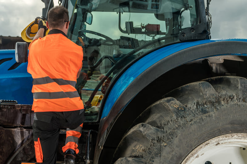 A farmer inspecting a stored tractor, checking for fluid leaks, battery condition, and tire pressure.