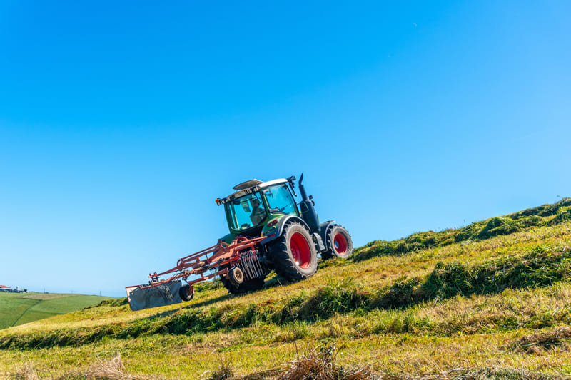 A farm tractor operating cautiously on a grassy slope, maintaining balance and traction to prevent rollover.