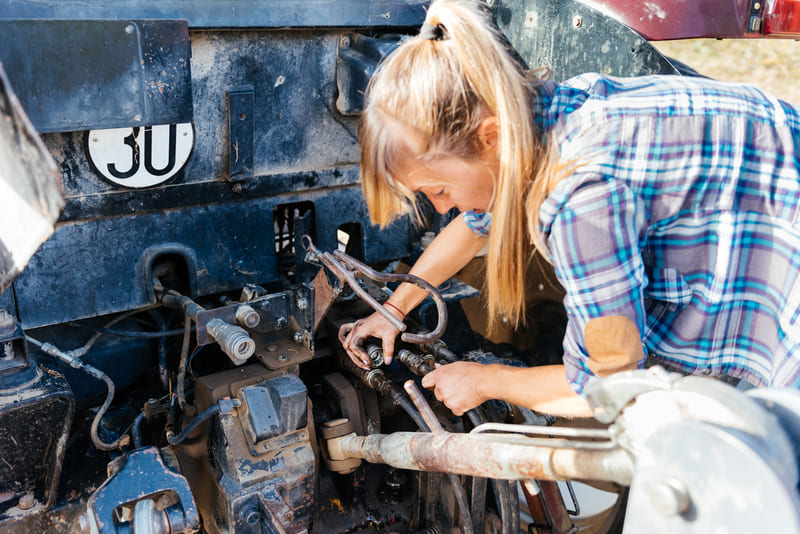 A farmer performing routine maintenance on a farm tractor, checking fluids, inspecting tires, and cleaning filters.
