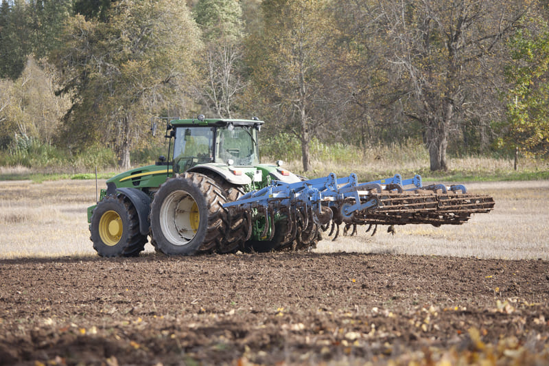 A farm tractor connected to multiple implements, including a rear-mounted tiller and a front-end loader, ready for multi-task fieldwork.