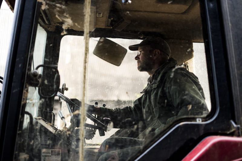 Man sitting inside winter tractor cab, dressed warmly, operating machinery in snowy landscape.